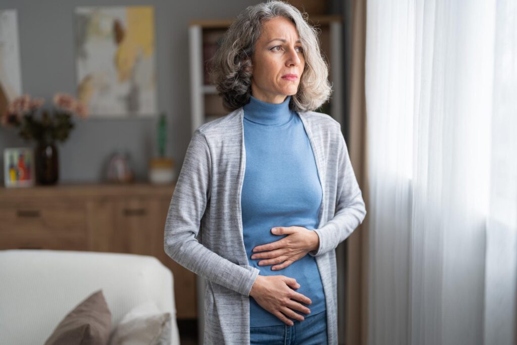Mujer latina de más de 40 años tocando su abdomen con expresión pensativa, reflejando barriga hinchada en la menopausia e incomodidad corporal.